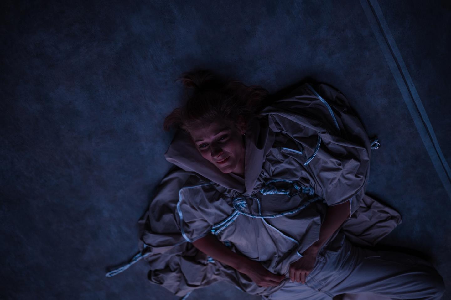 Portret z góry aktorki w ciemnoszarym kostiumie leżącej na podłodze i wpatrzonej w sufit.