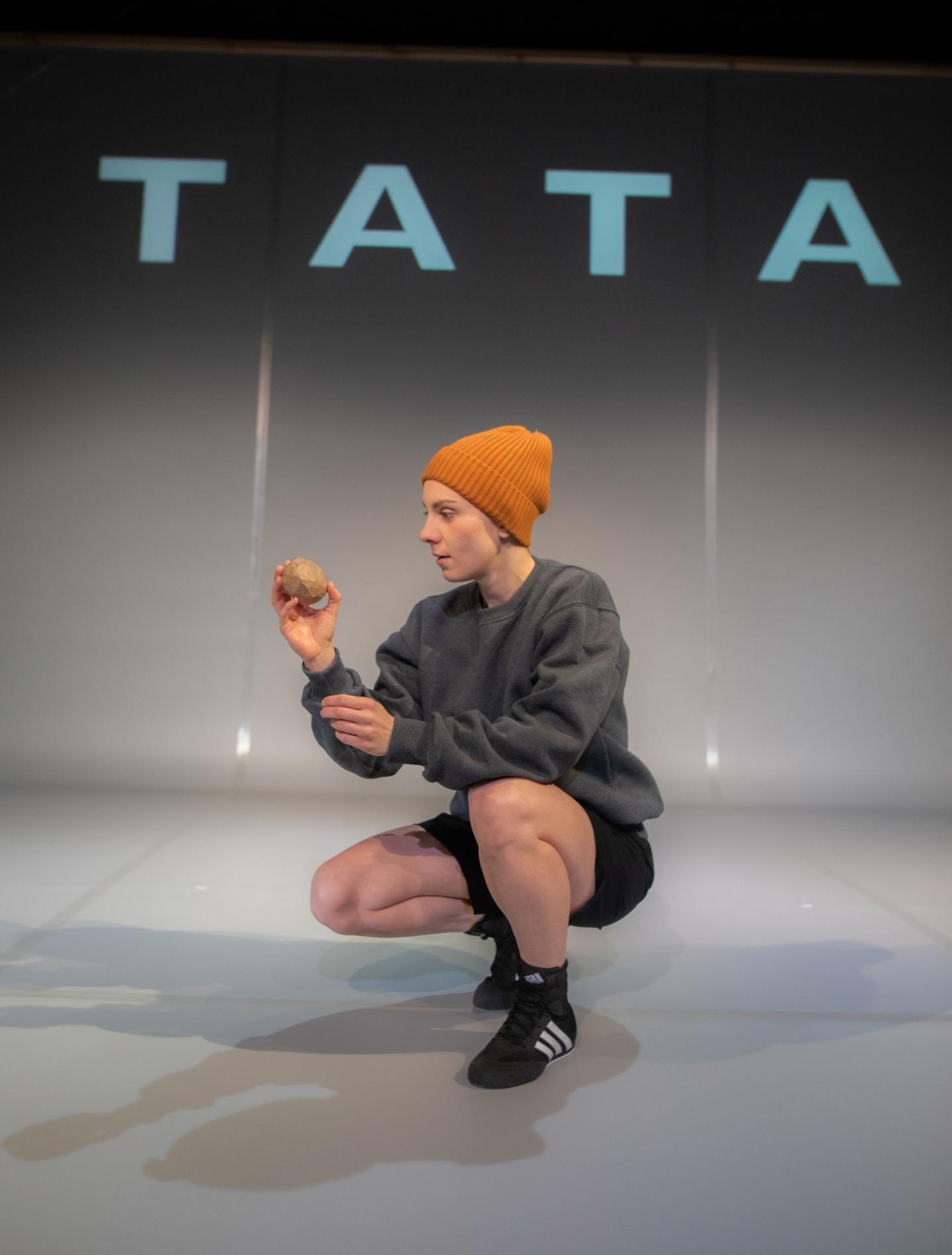 Kucająca kobieta w pomarańczowej czapce. Za nią na ścianie wyświetlony jest napis "Tata"