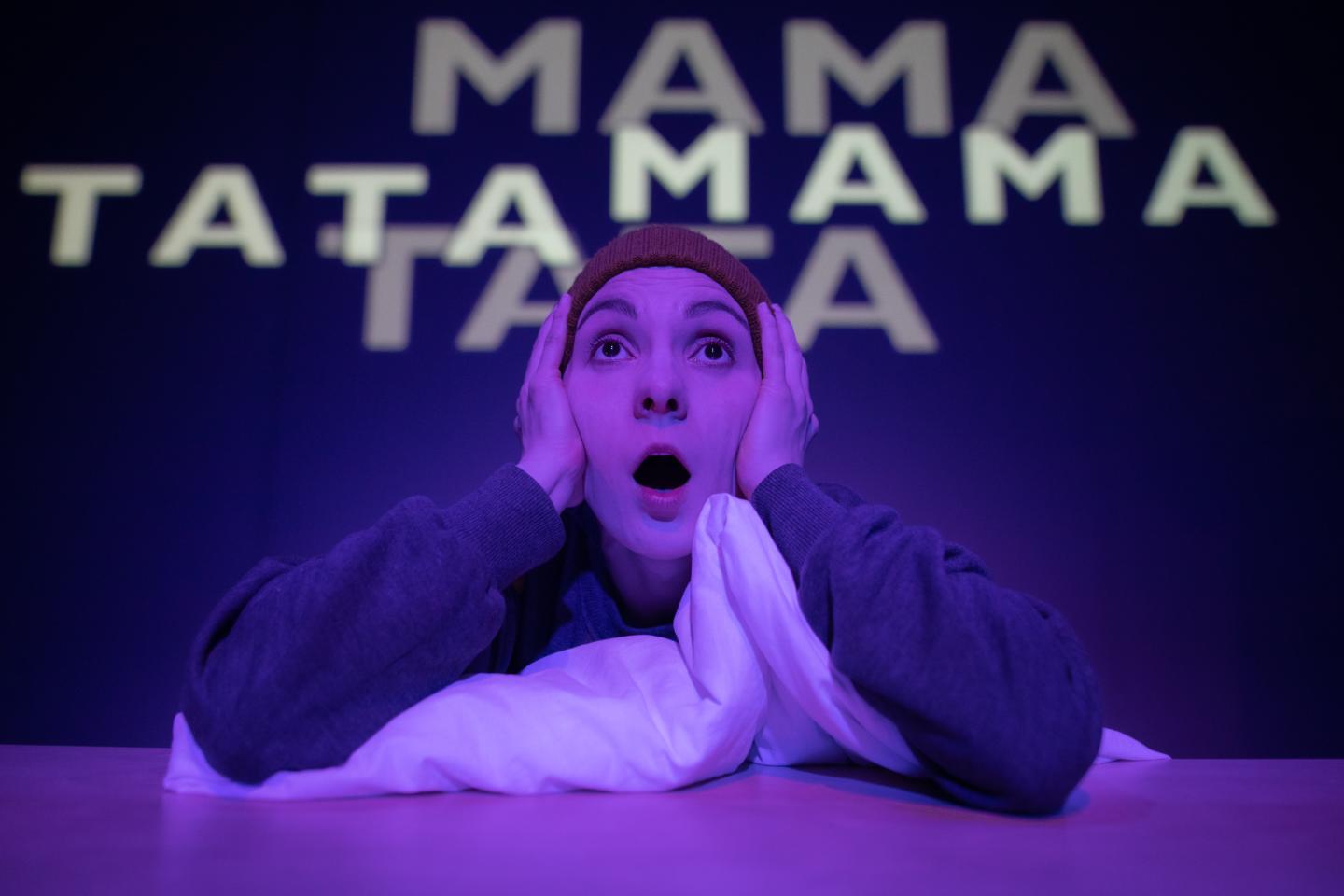 W fioletowym świetle zszokowana kobieta w pomarańczowej czapce patrzy przed siebie. Za nią na ścianie wyświetlone są napisy "Mama" i "Tata"