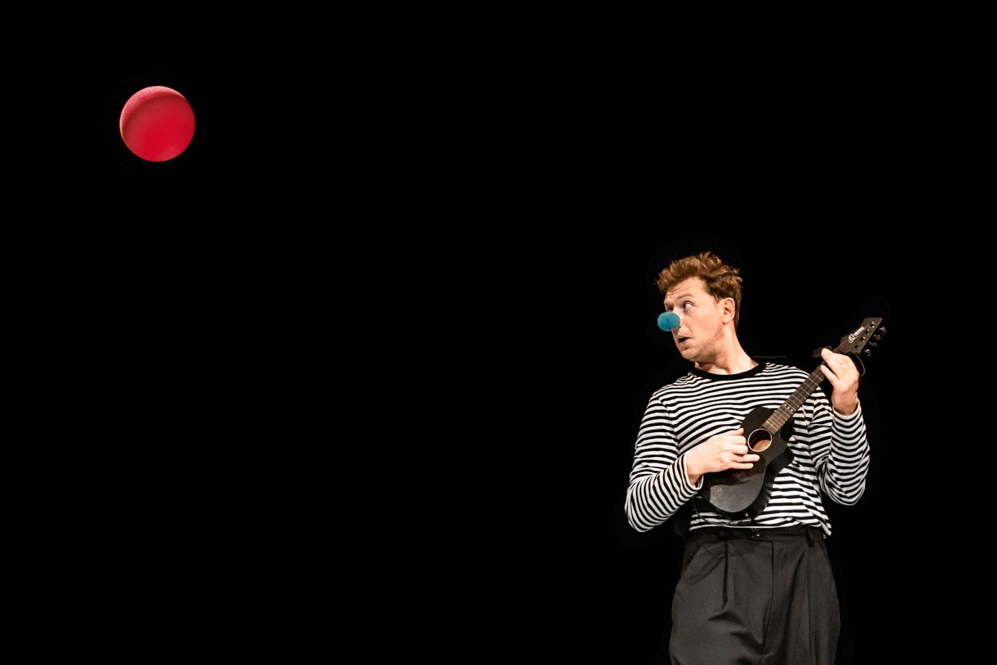 Na ciemnym tle mężczyzna z niebieską kulką na nosie gra na ukulele. Patrzy w prawo na wysoko znajdująca się czerwoną kulkę.