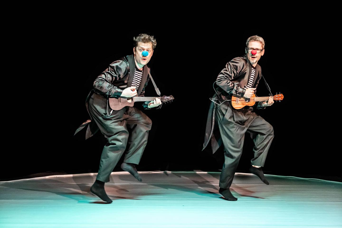 Dwóch mężczyzn w czarnych strojach i kolorowymi kulkami na nosach grają na ukulele.