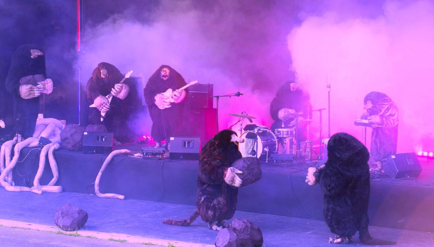 W fioletowym świetle i unoszącym się dymie, grupa muzyków w strojach kretów gra koncert.