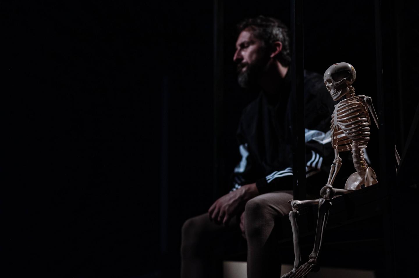 Mężczyzny siedzący w ciemnym oświetleniu, obok niego w tej samej pozie siedzi mały szkielet człowieka.
