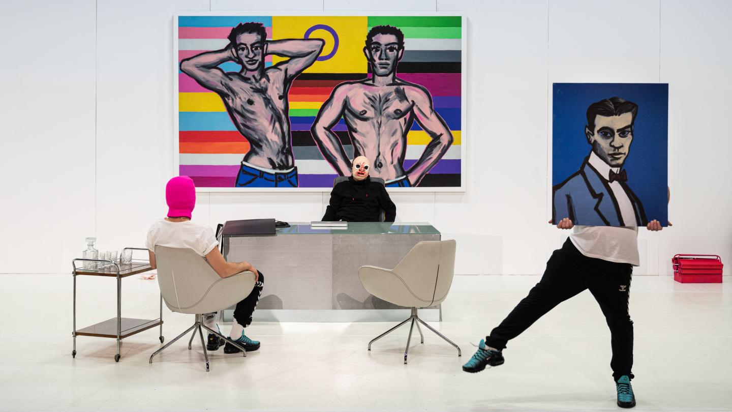 Nowoczesne, jasne wnętrze przypominające galerię sztuki lub minimalistyczne biuro. Na ścianie wisi duży, kolorowy obraz przedstawiający dwóch półnagich mężczyzn na tle poziomych pasów w barwach tęczy. Przy szklanym biurku siedzi osoba w czarnym stroju i masce klauna. Naprzeciwko niej, na jasnym fotelu, siedzi osoba w białej koszulce i różowej kominiarce. Po prawej stronie trzecia osoba stoi w rozkroku i trzyma przed twarzą obraz z portretem mężczyzny w garniturze. 