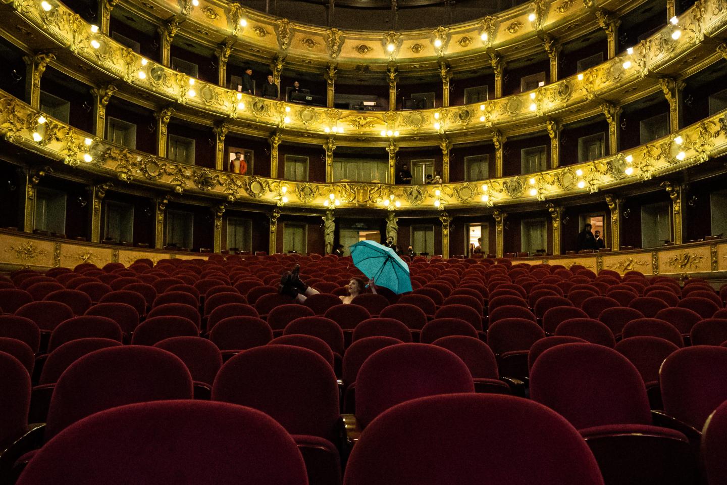 Widownia sali teatralnej. W tle złote balkony. Na środku rzędów foteli siedzi kobieta ubrana jedynie w ciemne buty. W ręku trzyma niebieską parasolkę.