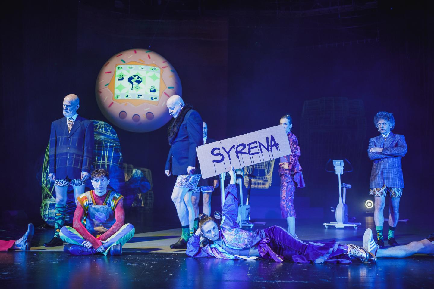 Grupa aktorów na scenie. Na pierwszym planie leży kobieta trzymająca transparent z napisem "syrena". Po jej prawej stronie siedzi mężczyzna ze skrzyżowanymi nogami. Na drugim planie znajdują się mężczyźni w marynarkach i krótkich spodenkach oraz kobieta we wzorzystym garniturze. W tle widać podwieszoną wielką kulę, na której wyświetlane jest różowe tamagotchi. 